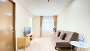 A modern living room with a sofa, a TV on a wooden stand, a small table with water bottles, and a fan. - Spacious 2Br Apartment At Grand Asia Afrika (Bandung)