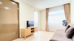 A living room with a TV on a wooden stand, a sofa, a window with curtains, and a door. - Spacious 2Br Apartment At Grand Asia Afrika (Bandung)