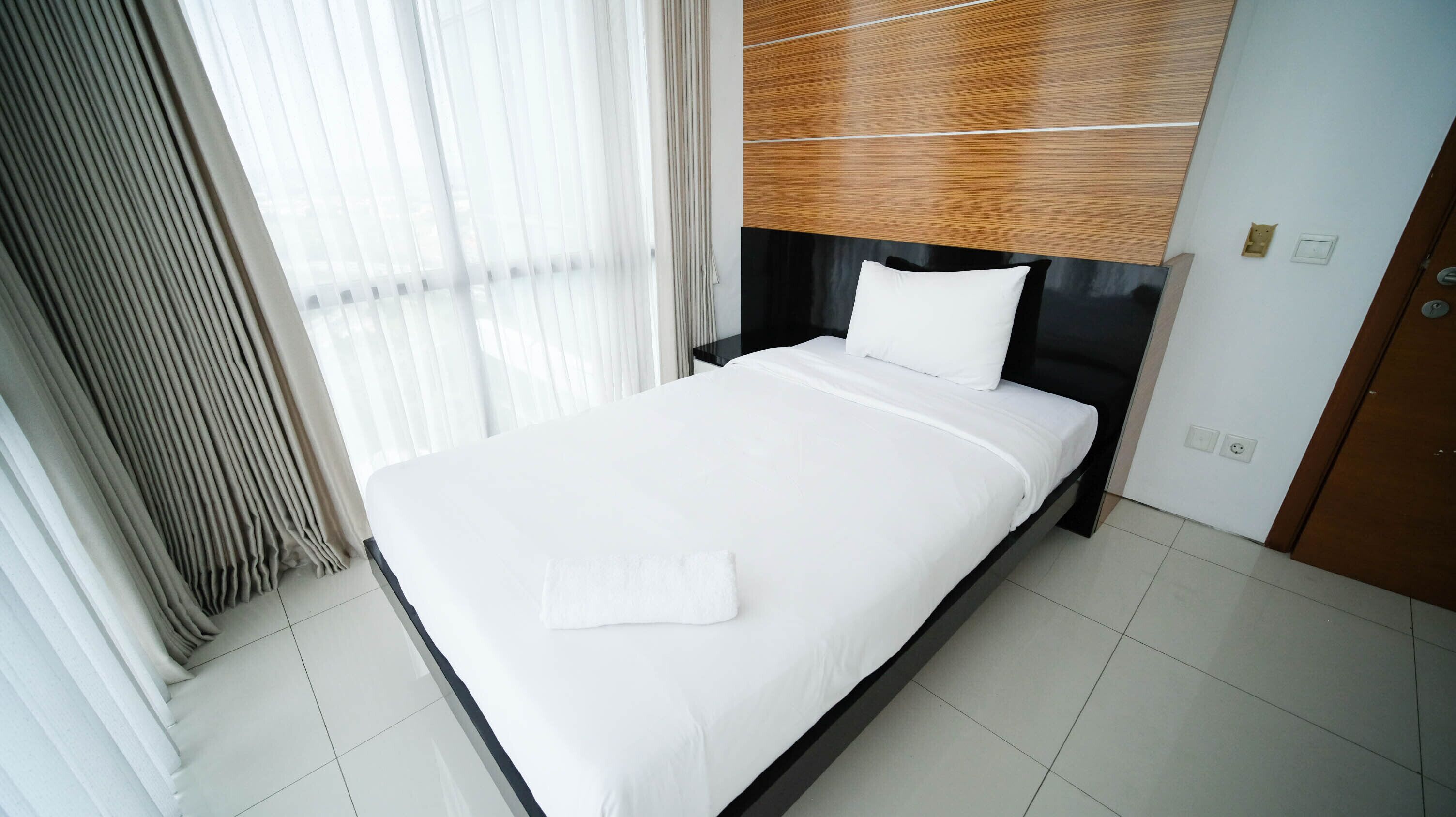 Ein modernes Hotelzimmer mit einem großen Bett, einem Holz-Kopfteil und Blick durch transparente Vorhänge.