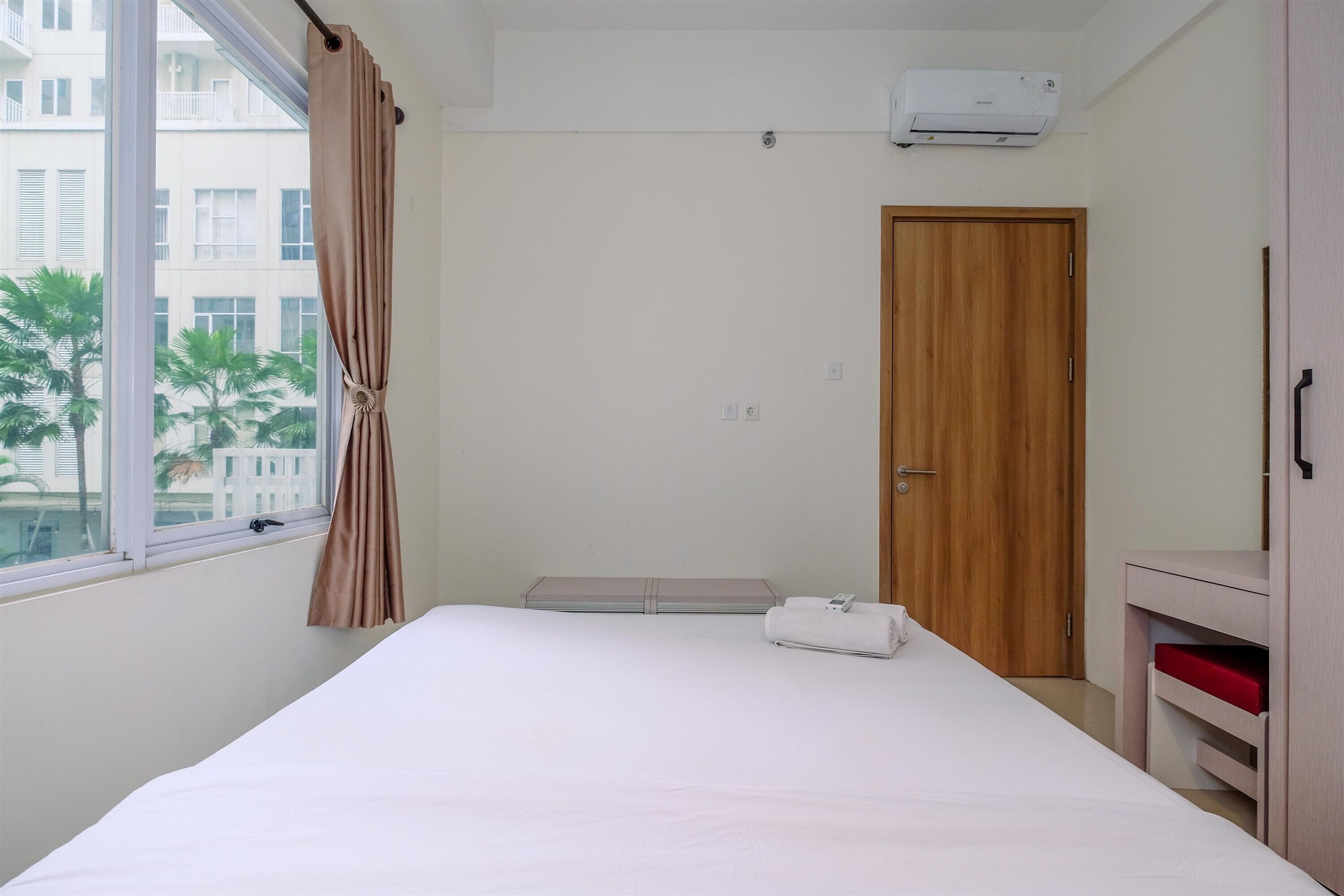 A bedroom with a bed, a window with curtains, a wooden door, and a small table with a red item.
