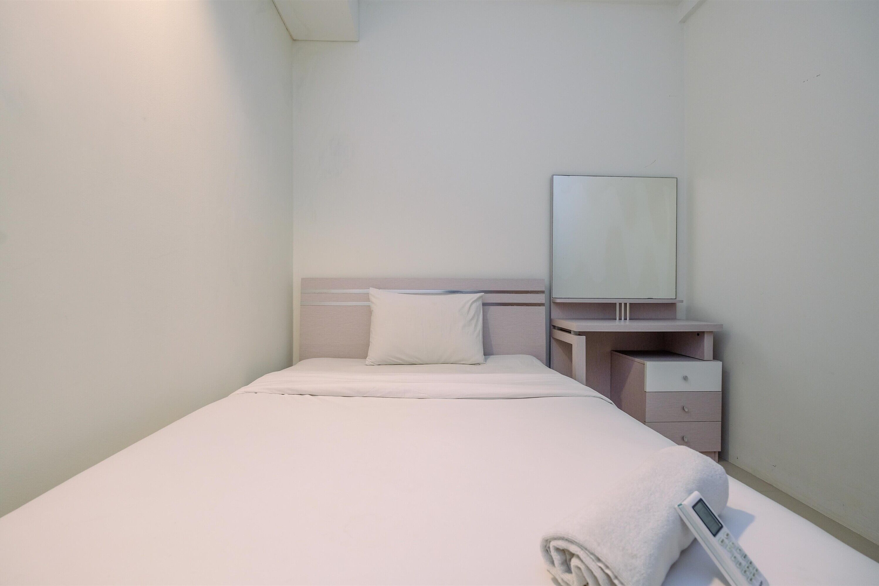 A neatly made bed with a white blanket and pillow, a bedside table with a lamp, and a small drawer unit.