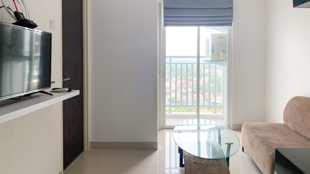 A modern living room with a flat-screen TV, a glass-top coffee table, a sofa, and a balcony with a view of the city. - Homey And Simply 2Br At Serpong Garden Apartment (Cisauk)