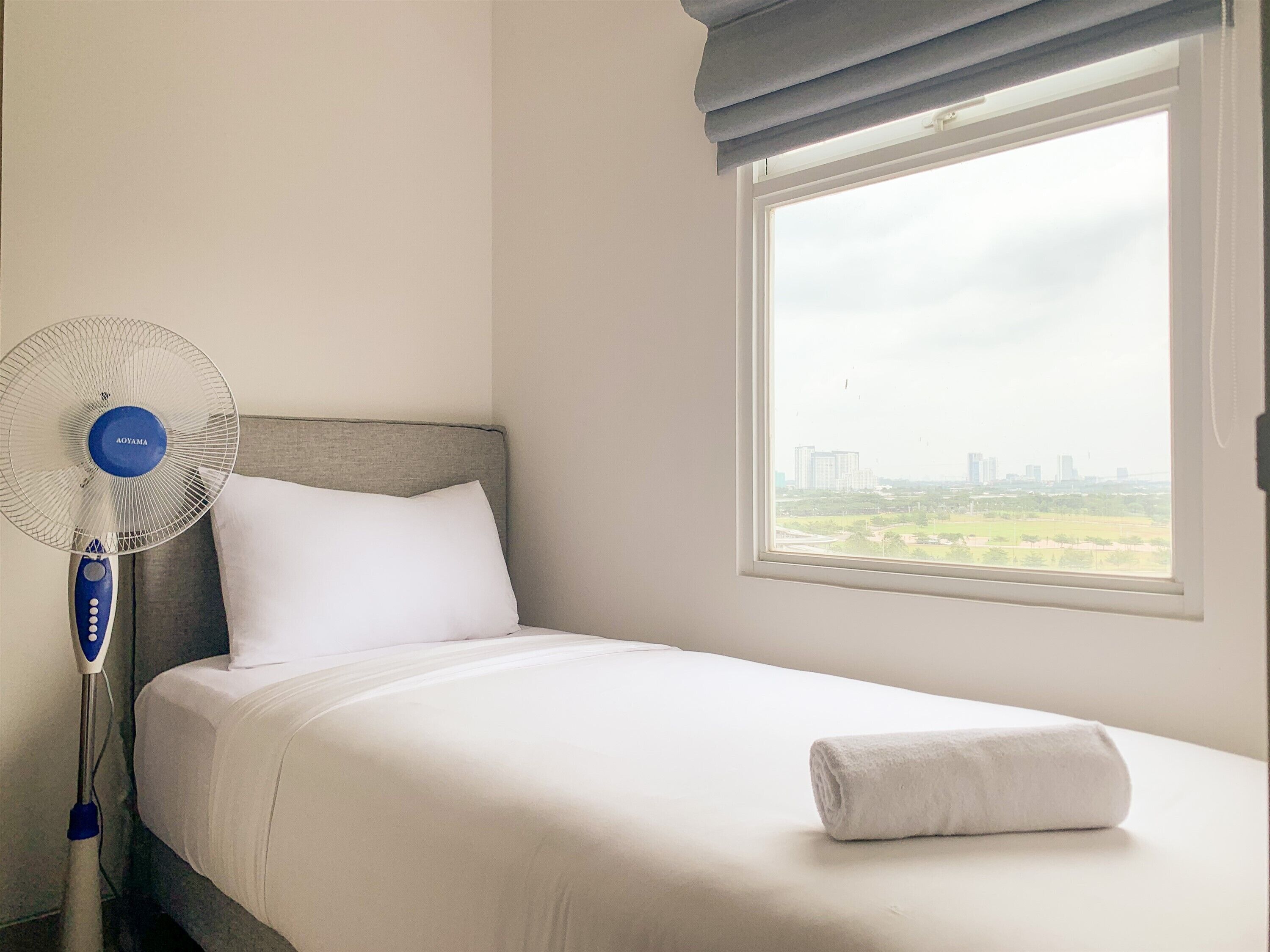 A hotel room with a bed, a standing fan, a window with a view of buildings, and a towel on the bed.