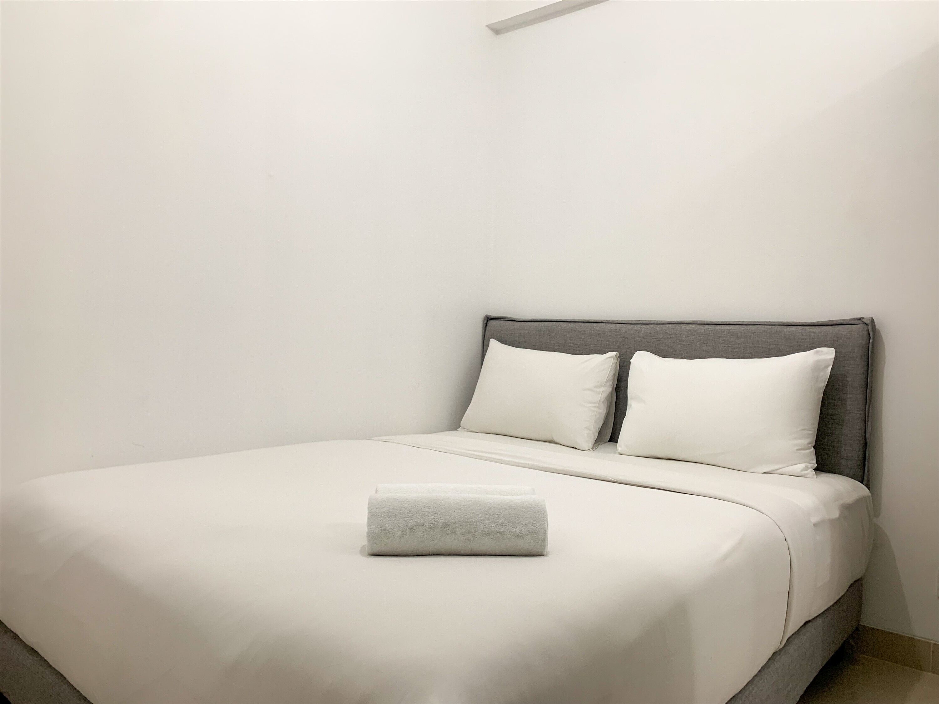 A neatly made bed with white bedding and pillows, a grey headboard, and a rolled white towel on the bed.