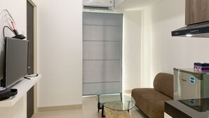 A modern living room with a flat-screen TV, a glass coffee table, a brown sofa, and a compact refrigerator. - Homey And Simply 2Br At Serpong Garden Apartment (Cisauk)