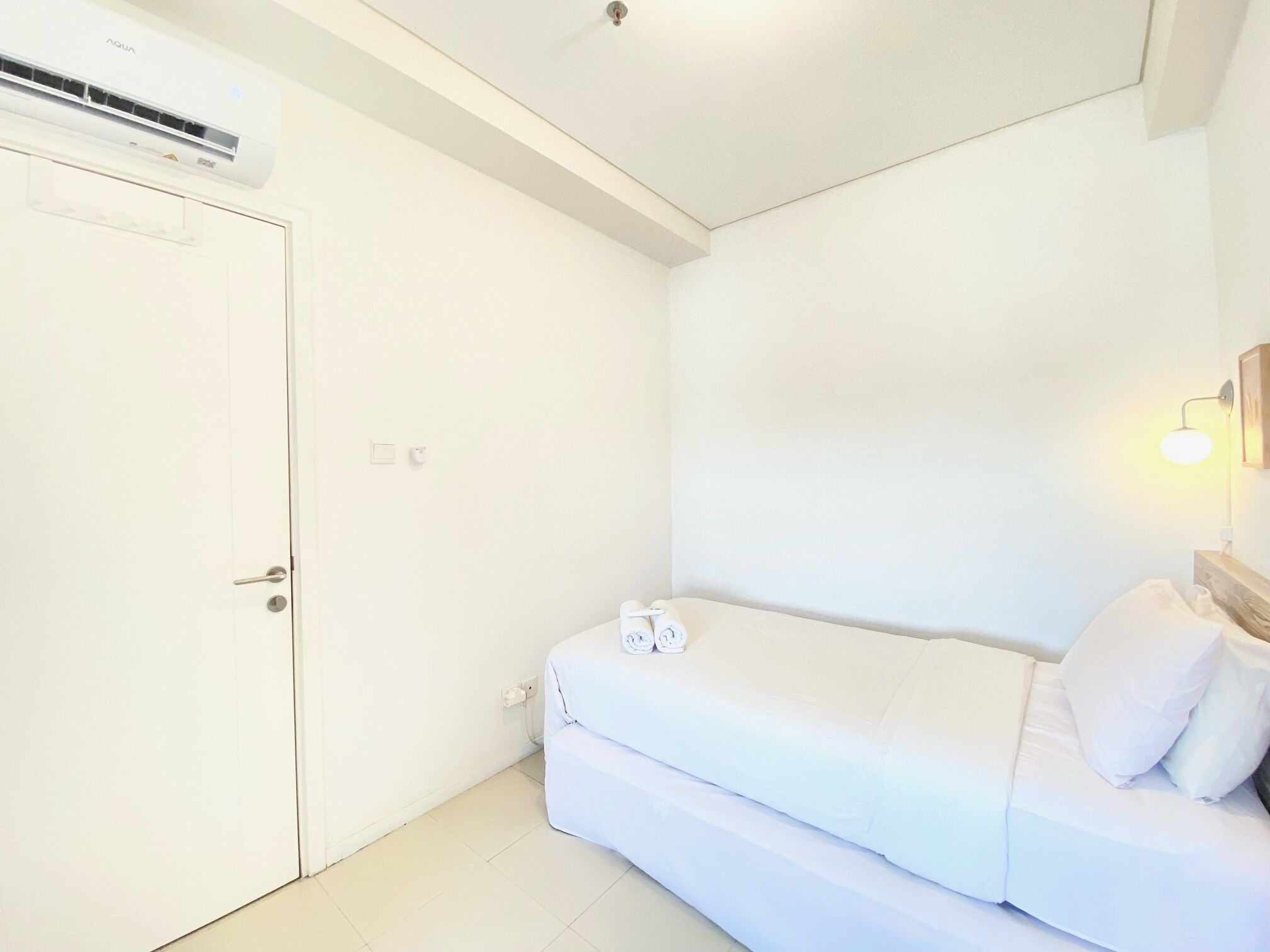 A small, white room with a bed, a door, and an air conditioning unit.