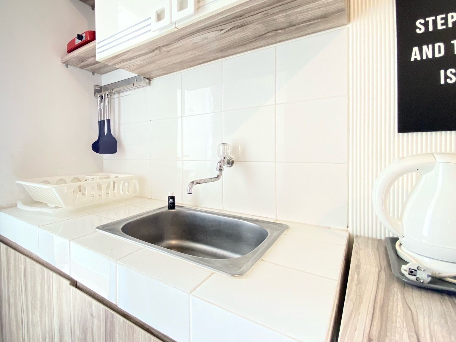 A kitchen with a stainless steel sink, white tiles, a wooden countertop, and a white kettle on the right.
