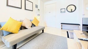 Un salon moderne avec un canapé gris, des coussins jaunes et beiges, une table basse, un téléviseur et une bibliothèque.