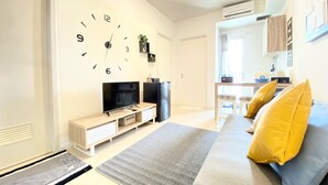 Un salon moderne avec une grande horloge murale, un téléviseur à écran plat, un canapé avec des coussins jaunes et, au fond, un coin cuisine.