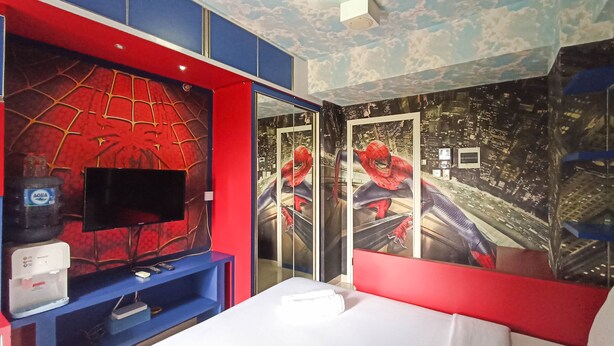 A hotel room with a large bed, a TV mounted on a red wall, and Spider-Man themed decorations.