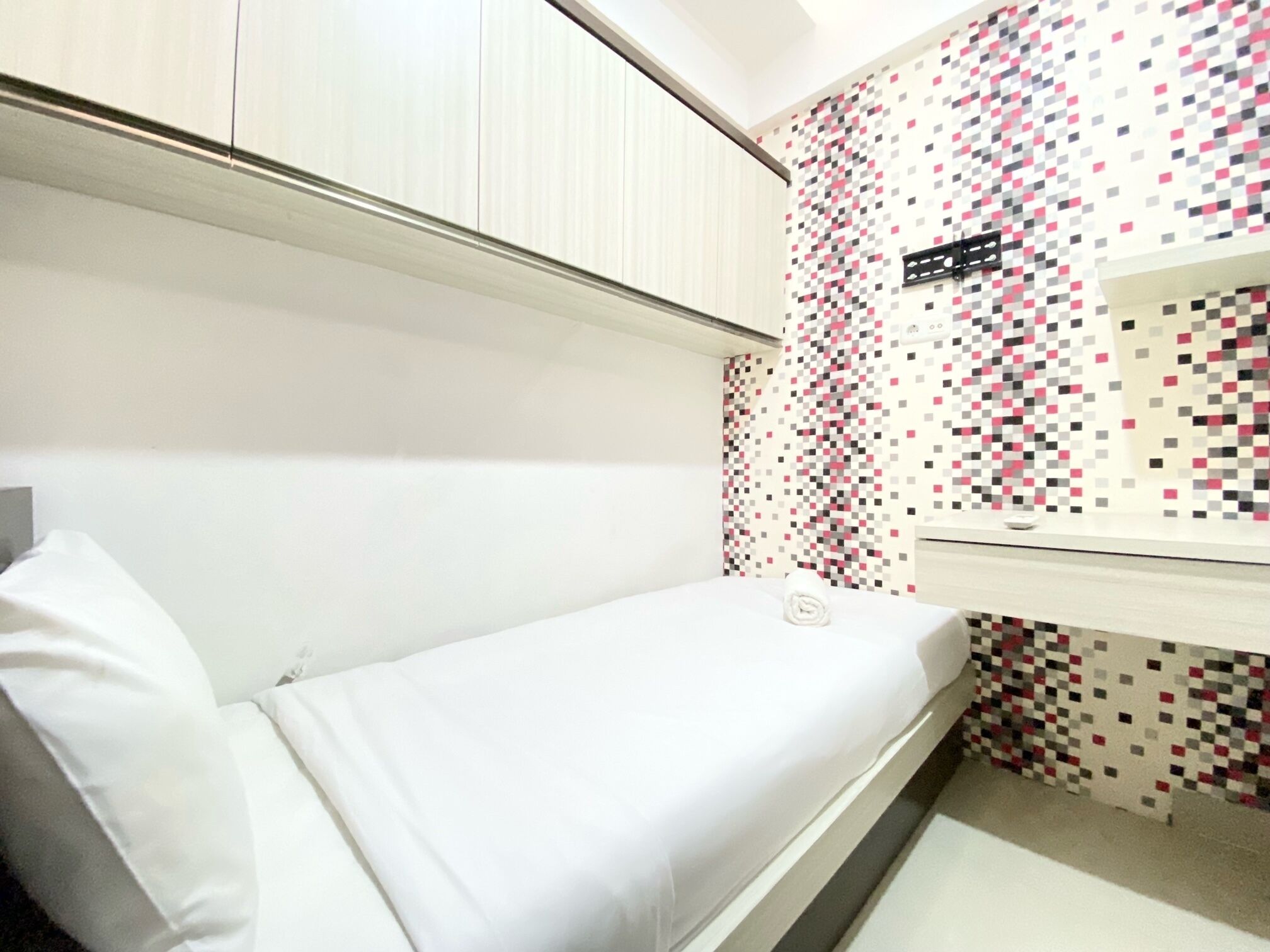 Une petite chambre avec un lit simple, un placard blanc et un mur orné d’un motif pixélisé.