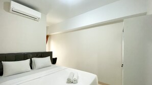 A hotel room with a bed, white linens, a headboard, a wall-mounted air conditioner, a door, and a nightstand with a lamp. - Elegant And Comfy 2Br Springlake Summarecon Bekasi Apartment Near Summarecon Mall (Bekasi)