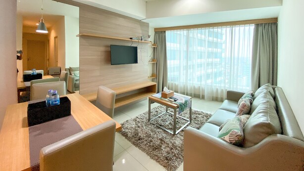 A modern living room with a flat-screen TV mounted on the wall, a wooden coffee table, a sofa, and a dining area with a table and chairs. - Fancy And Nice 3Br At Grand Kamala Lagoon Apartment (Bekasi)