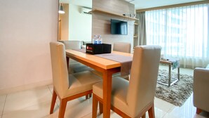 A modern hotel room with a wooden dining table, four chairs, a flat-screen TV mounted on the wall, and a large window with sheer curtains. - Fancy And Nice 3Br At Grand Kamala Lagoon Apartment (Bekasi)
