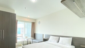 A hotel room with a bed, a bedside table, a wardrobe, and a window with curtains. - Enjoy Living Studio Room At High Floor Grand Kamala Lagoon Apartment (Bekasi)