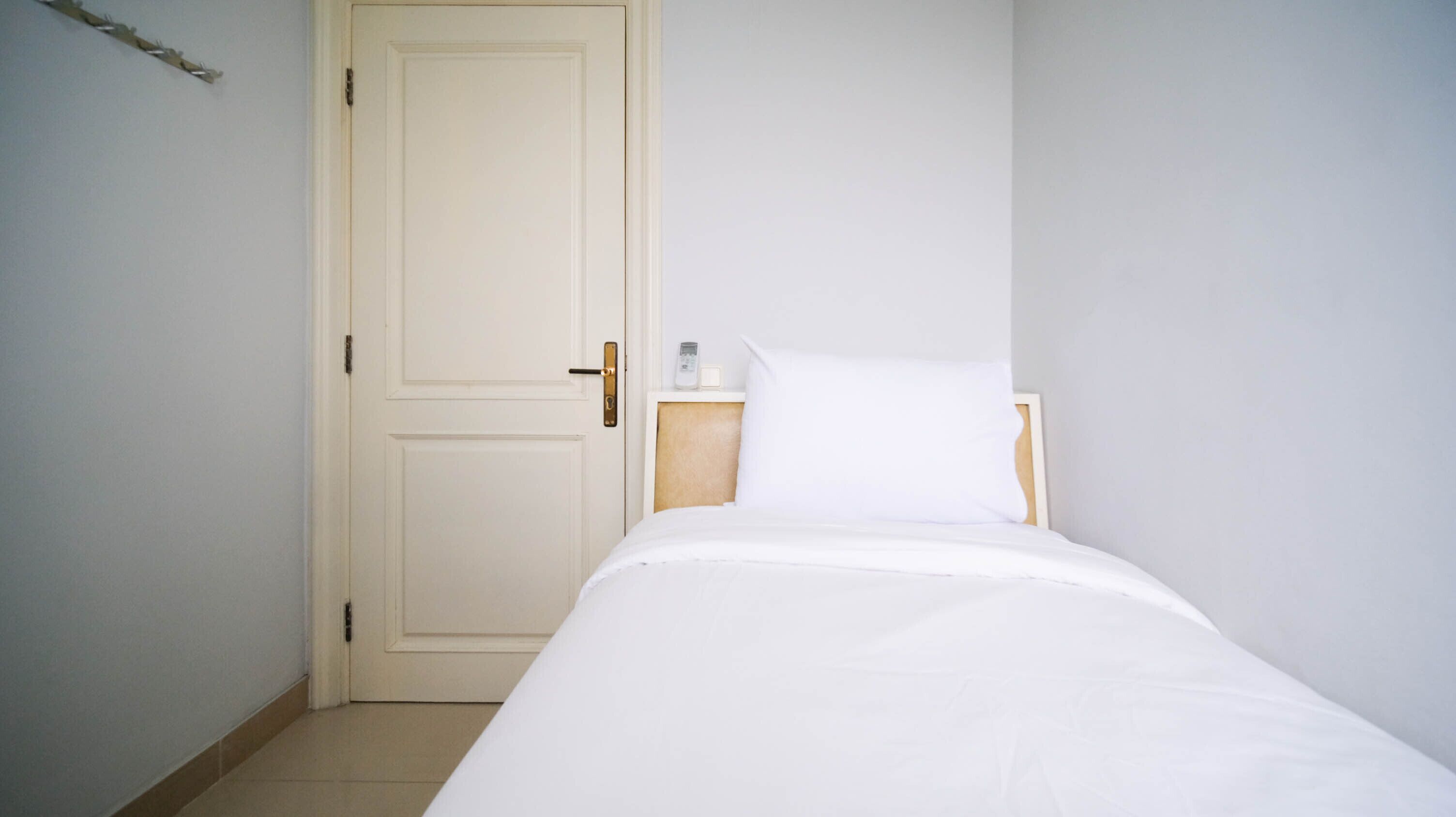 Una habitación pequeña y limpia con una cama individual, una mesita de noche de madera y una puerta blanca.