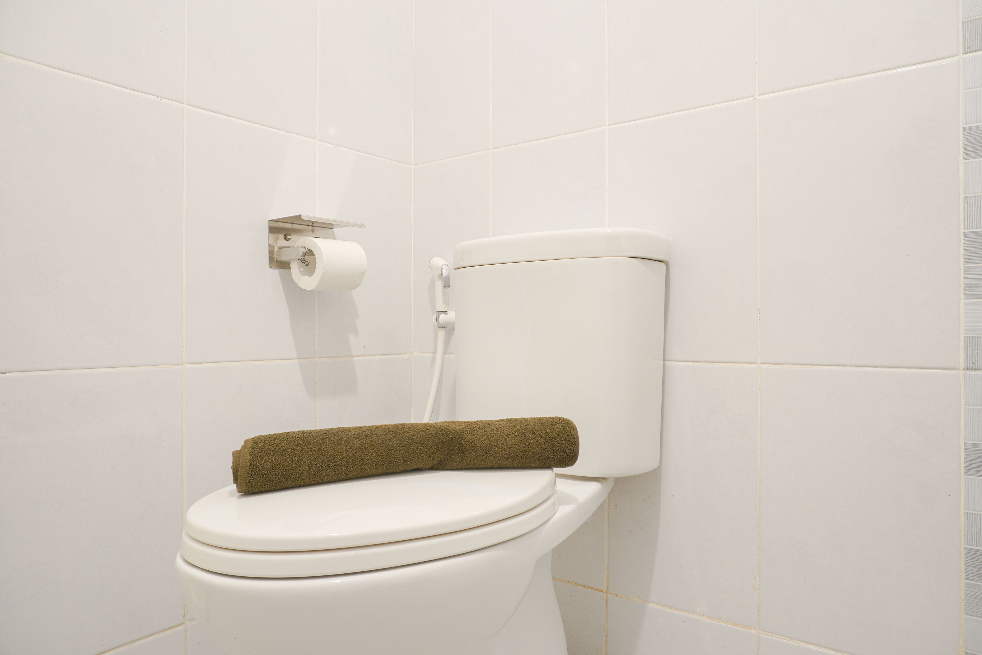 Des toilettes blanches avec une serviette marron posée sur le couvercle, un porte-rouleau de papier toilette fixé au mur et des murs recouverts de carreaux blancs.