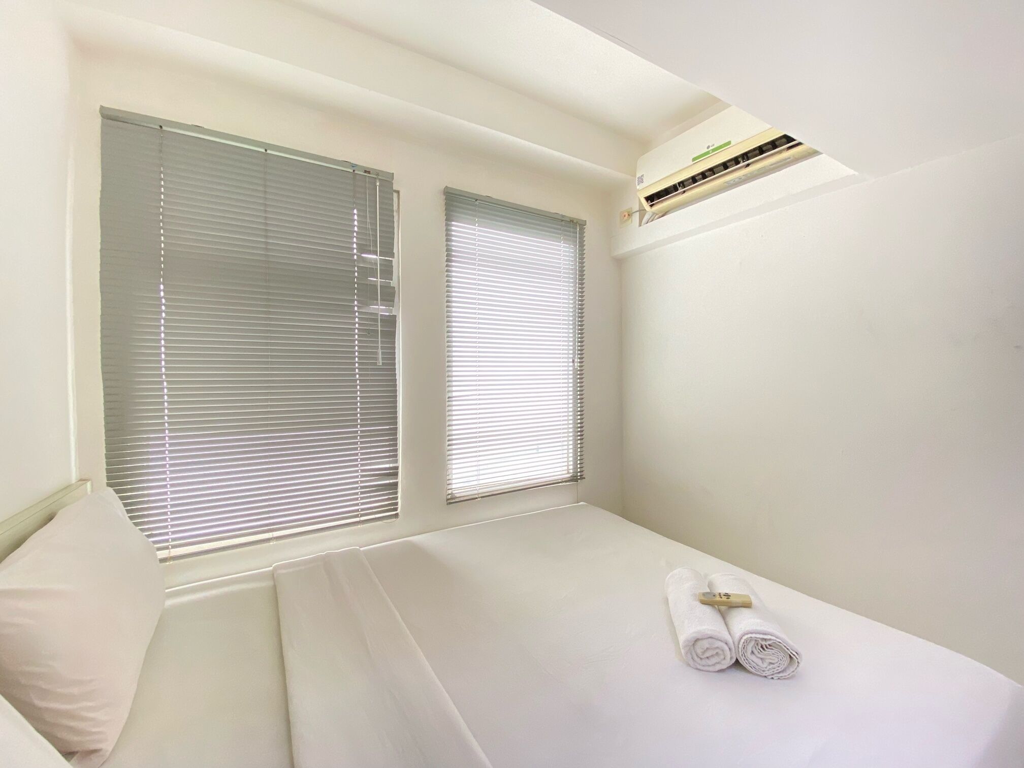 A small, neatly made bed with white linens and two rolled towels, a window with blinds, and an air conditioning unit on the wall.