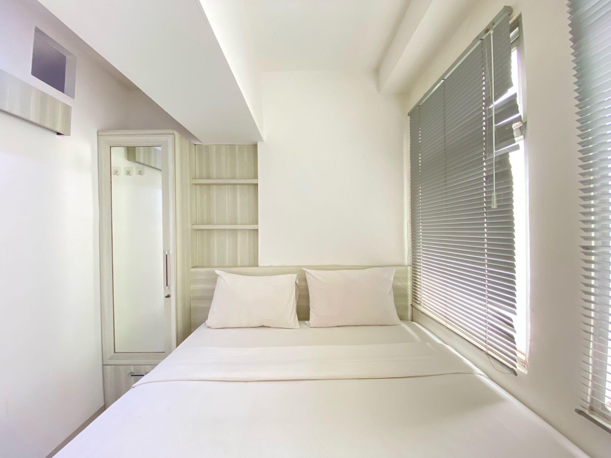 Une chambre à coucher avec un lit, deux oreillers, une fenêtre avec des stores, une porte et une petite étagère.