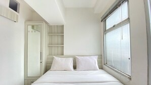 Een kleine, witte kamer met een enkel bed, een raam, een kast en een deur.