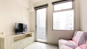 Een lichte, kleine kamer met een televisie op een houten kast, een bank met gekleurde kussens en twee ramen met uitzicht op gebouwen.