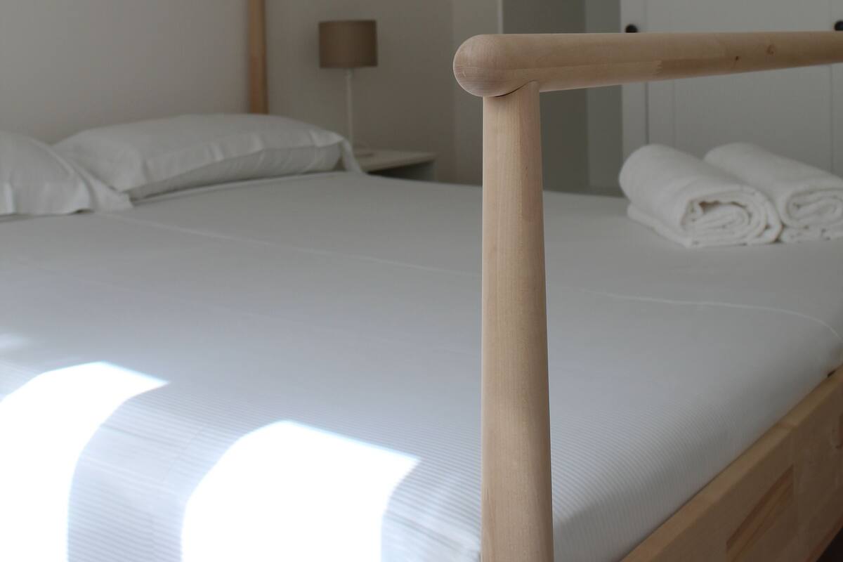 A neatly made bed with white linens, a wooden headboard, and a bedside lamp. There is a wardrobe with a mirror and a door leading to a bathroom.