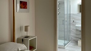 A bedroom with a bed, a bedside table with a lamp, a framed picture on the wall, and a door leading to a bathroom with a shower and sink. - Virgilio 82 - Penthouse With Terrace (Trapani)