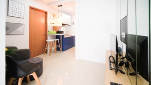 Un saló modern amb un sofà i una televisió. Hi ha una zona de cuina amb nevera, rentapelats i tamborets.