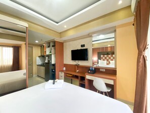 A hotel room with a bed, a desk, a TV, a mirror, and a bathroom. - Cozy Studio Room At Oasis Cikarang Apartment (Cikarang)
