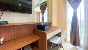 A modern hotel room with a wooden desk, a flat-screen TV, a window with curtains, and a ceiling-mounted air conditioner.