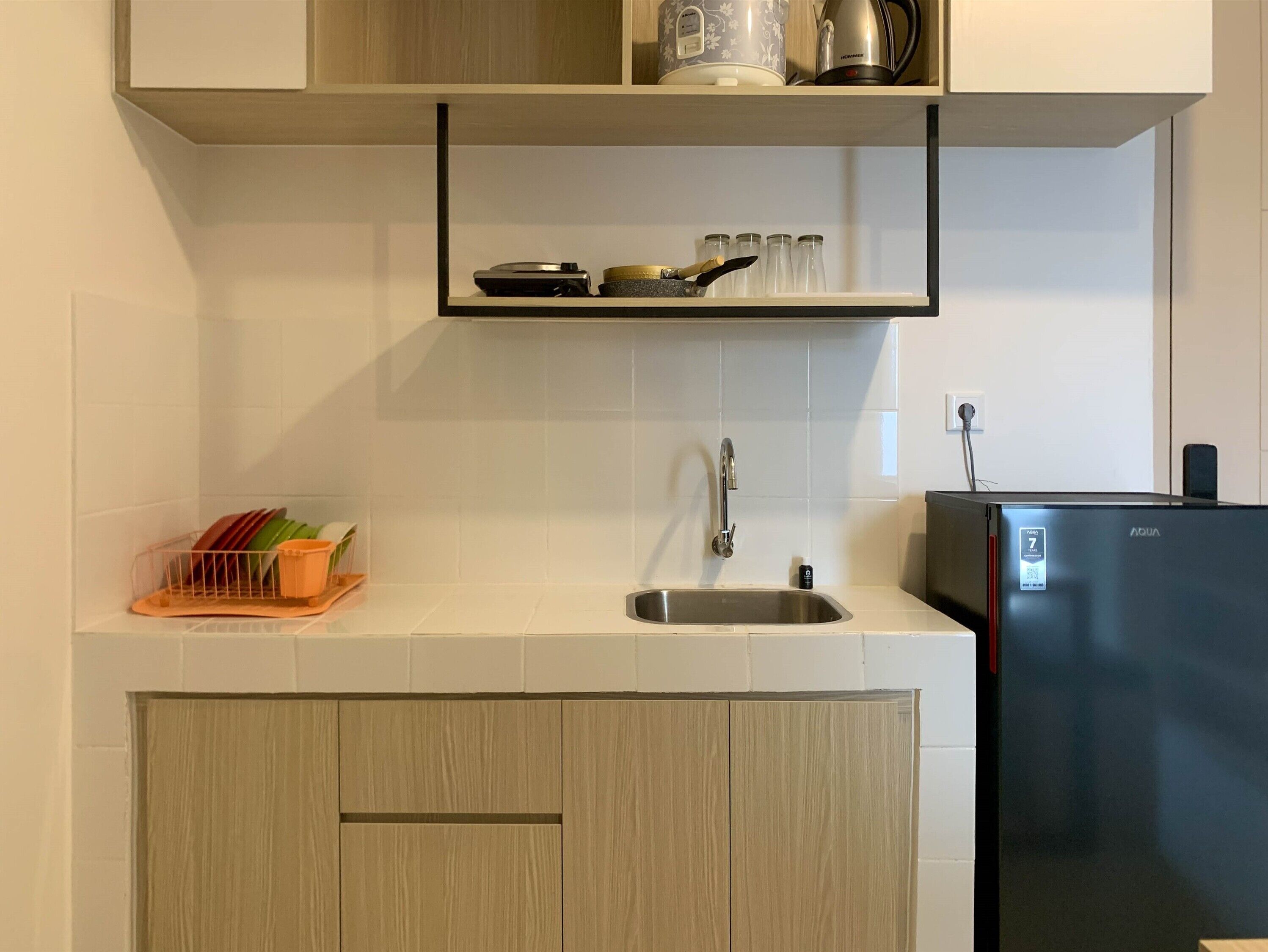 A kitchen with a stainless steel sink, a black refrigerator, and a shelf above the sink holding kitchenware.