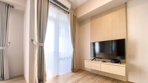 A modern living room with a flat-screen TV on a wooden cabinet, a sliding glass door with curtains, and a wooden floor. - Cozy And Simple Big Studio Vasanta Innopark Apartment (Cikarang)