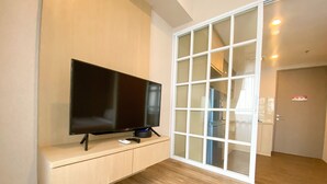 A modern living room with a flat-screen TV on a wooden console, a sliding glass door leading to a kitchen, and a wooden floor. - Cozy And Simple Big Studio Vasanta Innopark Apartment (Cikarang)