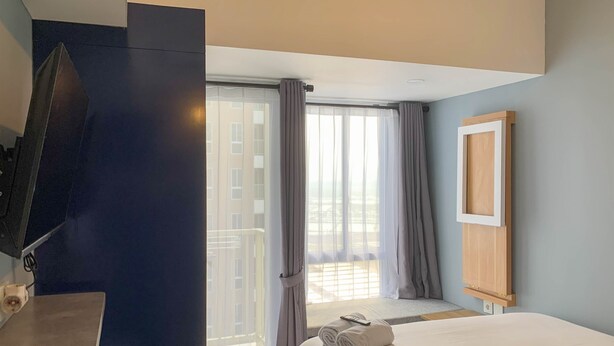 A hotel room with a bed, a TV mounted on a dark wall, a window with curtains, and a view of buildings outside. - Cozy Studio At 31Th Floor Tokyo Riverside Pik 2 Apartment (Teluknaga)