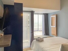 A hotel room with a bed, a TV mounted on a dark wall, a window with curtains, and a view of buildings outside.