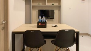 Una habitación pequeña y moderna con un escritorio de madera, dos sillas negras, un televisor montado en la pared y un estante con botellas de agua y una bolsa de viaje.