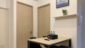 Una habitación de hotel con una puerta de madera, una mesita pequeña con una silla negra, una estantería con una planta y una bandeja con botellas de agua.
