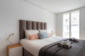 Ein modernes Schlafzimmer mit einem großen Bett, einer Kopfteil, Nachttischlampen und Blick durch ein Fenster.