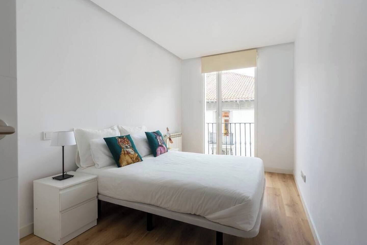 A bedroom with a bed, nightstand, lamp, and a view of the outside.