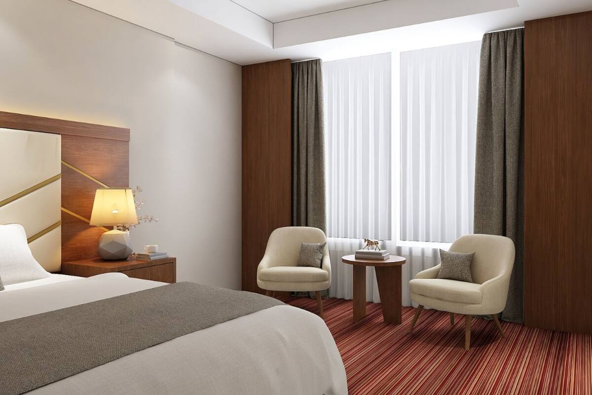 Quarto de hotel com uma cama grande, duas poltronas, uma mesinha, uma luminária e vista para o exterior através de cortinas leves.