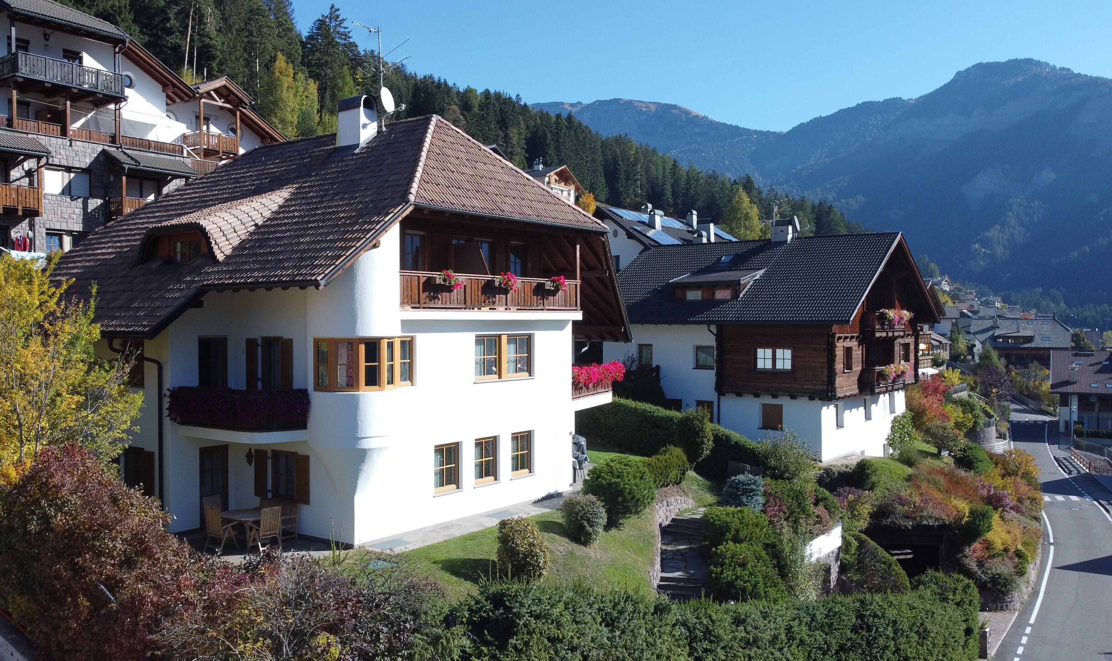 A traditional chalet-style building with a balcony, surrounded by lush greenery and mountains in the background.