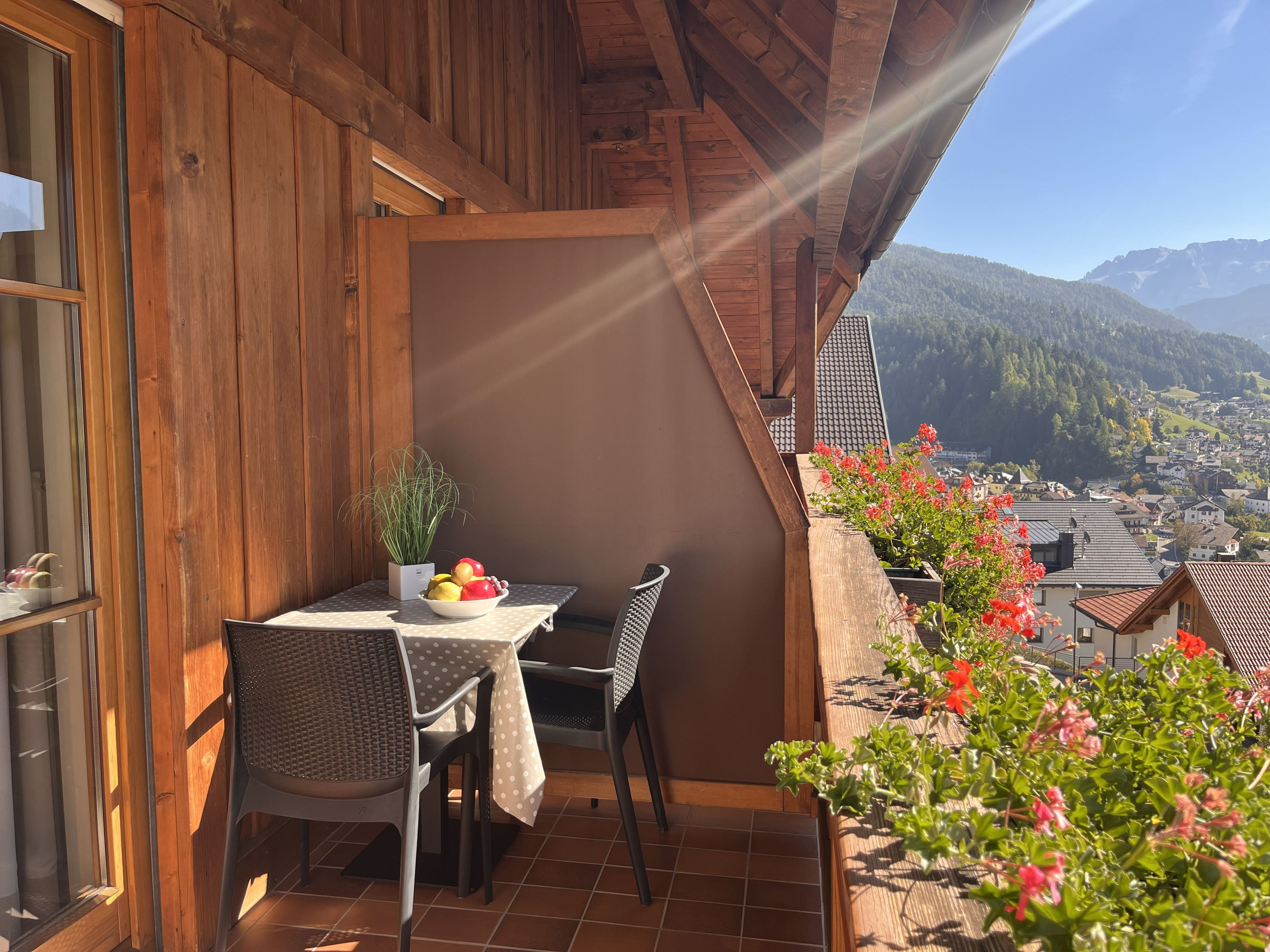 A wooden balcony with a table and chairs, a bowl of fruit, and a view of a town and mountains.