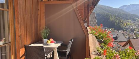 A wooden balcony with a table and chairs, a bowl of fruit, and a view of a town and mountains.