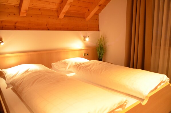 Un lit double avec du linge de lit blanc, dans une pièce dont le plafond est orné de poutres en bois et qui est éclairée par un luminaire fixé au mur.