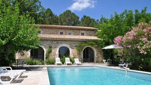 A stone building with a swimming pool, surrounded by greenery and white lounge chairs.