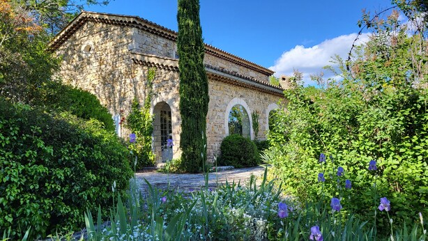 A stone building with arched windows and a tiled roof, surrounded by lush greenery and flowering plants.