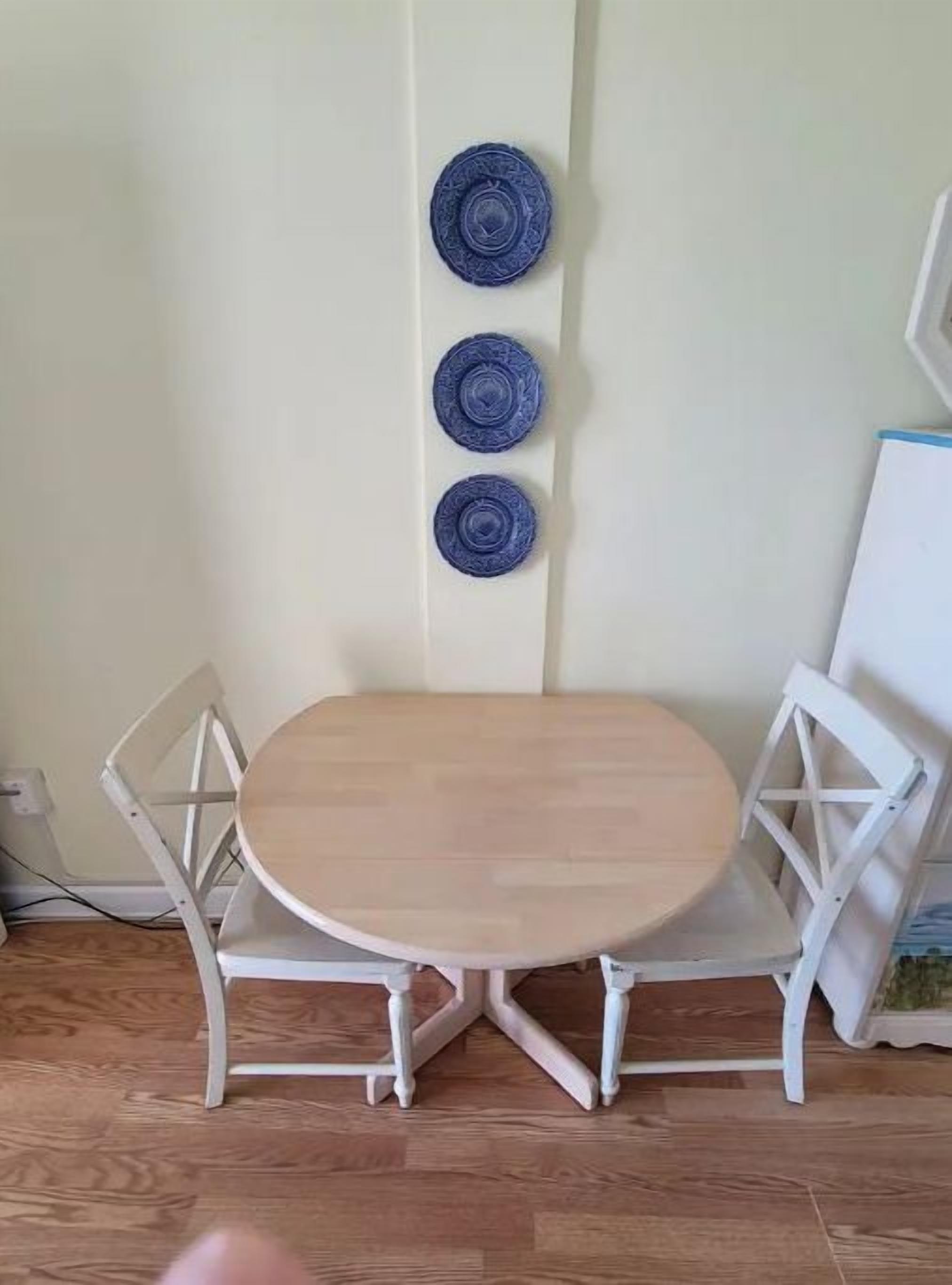 Un pequeño comedor con una mesa redonda y dos sillas blancas. Encima de la mesa, hay tres platos decorativos azules en la pared.