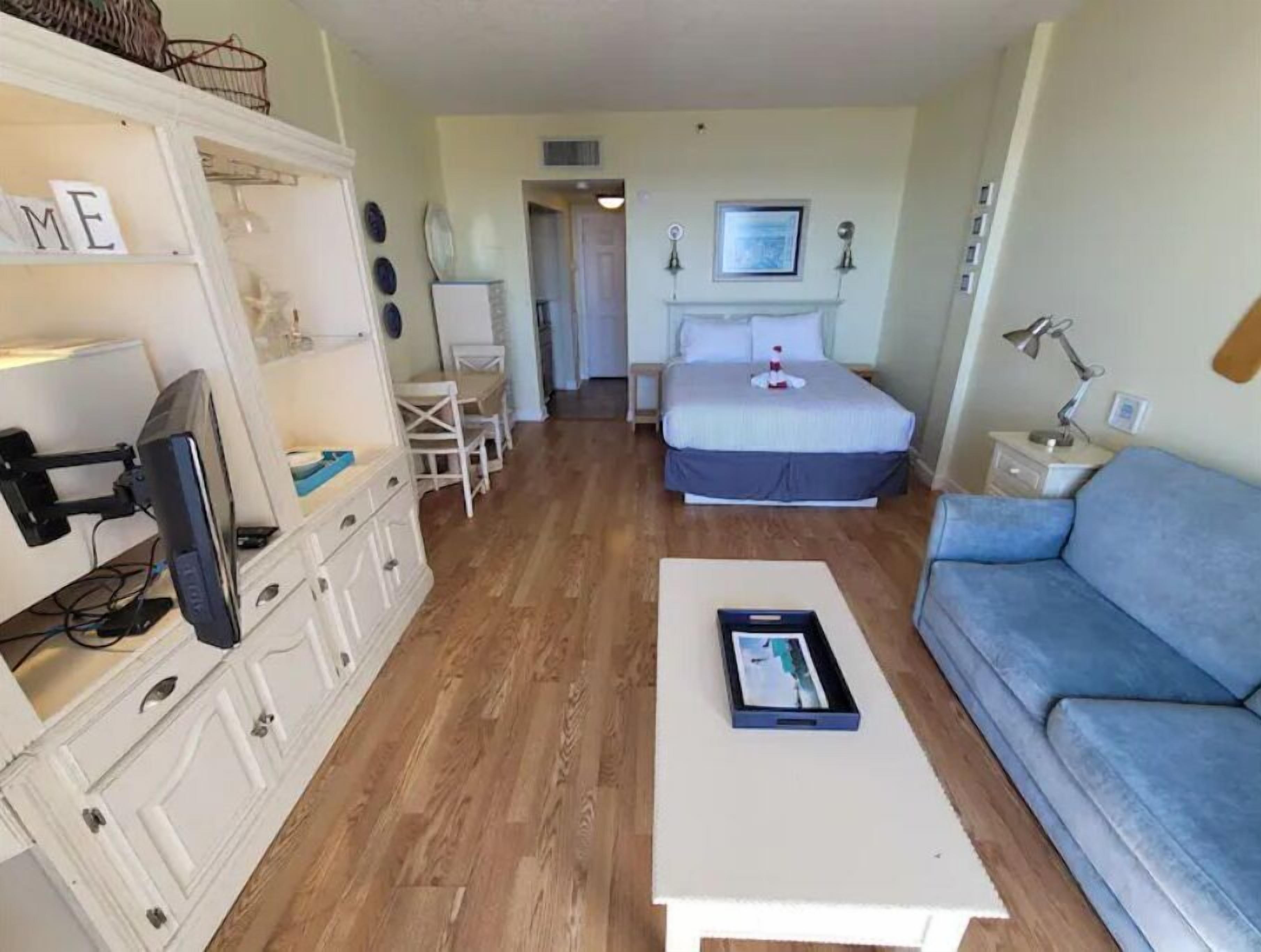 Ein kleines Zimmer mit einem Bett, einem blauen Sofa, einem weißen Couchtisch, einem Schreibtisch mit Computer und einer kleinen Essecke.