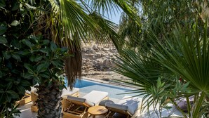 Ein Poolbereich mit Liegestühlen und einem Holztisch, umgeben von tropischen Pflanzen und einer felsigen Landschaft im Hintergrund.