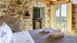 Ein Zimmer mit Steinwänden, einer Holzbalkendecke, einem Bett mit weißen Leinen und Blick durch ein Fenster.